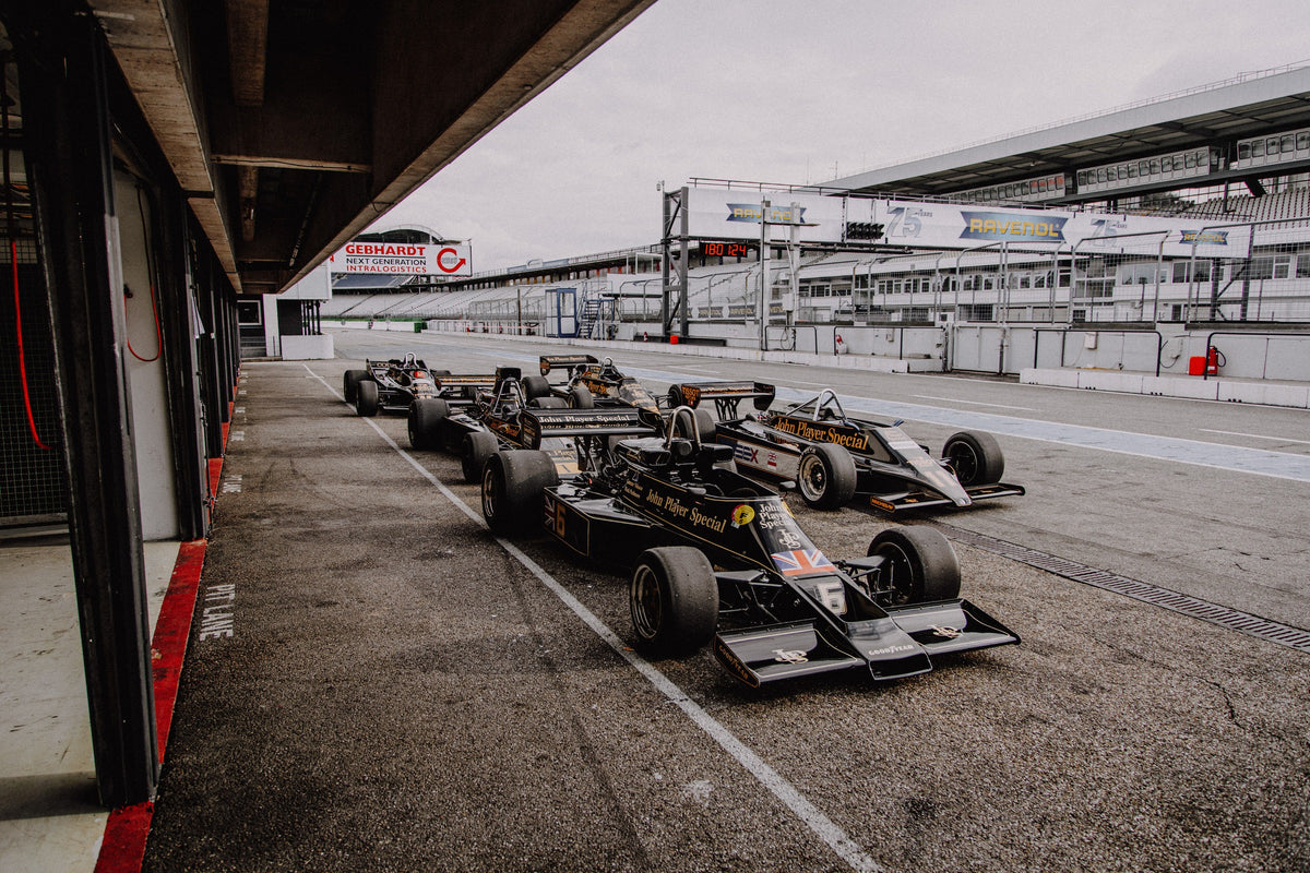 Mehrere historische Lotus-Rennwagen rollen in enger Formation an der Boxengasse entlang; der Blick von oben zeigt die Staffelung. Originalmotiv „Trackside Moment“ im Querformat.