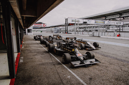 Mehrere historische Lotus-Rennwagen rollen in enger Formation an der Boxengasse entlang; der Blick von oben zeigt die Staffelung. Originalmotiv „Trackside Moment“ im Querformat.
