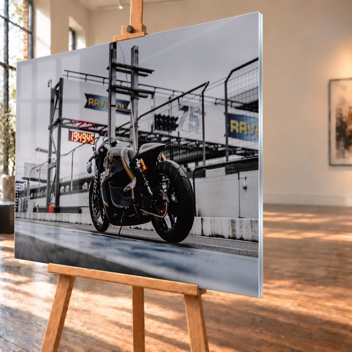 Als Acrylglasbild visualisiertes Motorradmotiv in schräger Ansicht auf einer Staffelei, mit schmaler rechter Kante und Fensterreflexionen auf der Bildfläche. Zu sehen ist der schwarz-goldene Lotus C-01 von schräg hinten vor einer Boxenanlage.