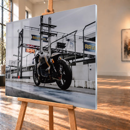 Als Acrylglasbild visualisiertes Motorradmotiv in schräger Ansicht auf einer Staffelei, mit schmaler rechter Kante und Fensterreflexionen auf der Bildfläche. Zu sehen ist der schwarz-goldene Lotus C-01 von schräg hinten vor einer Boxenanlage.