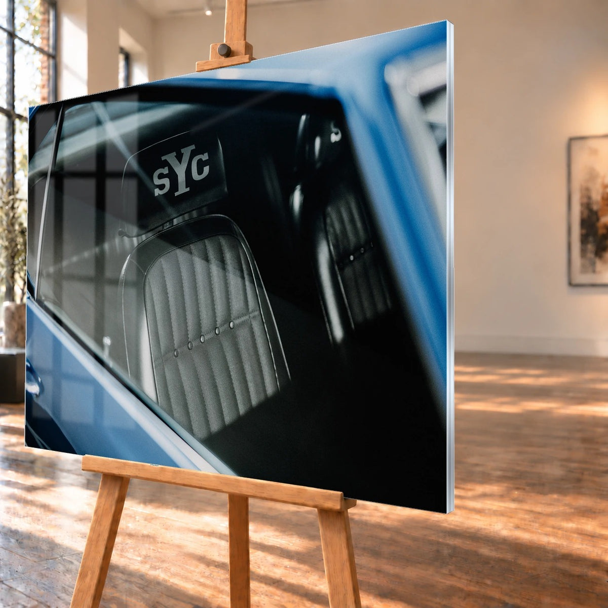 Die Innenraum-Nahaufnahme des blauen Camaro Yenko zeigt den hellen Sitz mit SYC-Schriftzug, eingefasst von Fenster und Karosserie. Motiv „Hero Moment 2“ als Acrylglas-Wandbild, schräg von der Seite auf einer Holzstaffelei.