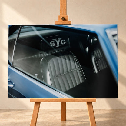 Die Innenraum-Nahaufnahme des blauen Camaro Yenko zeigt den hellen Sitz mit SYC-Schriftzug, eingefasst von Fenster und Karosserie. Motiv „Hero Moment 2“ als Alu gebürstet-Wandbild, frontal auf einer Holzstaffelei.