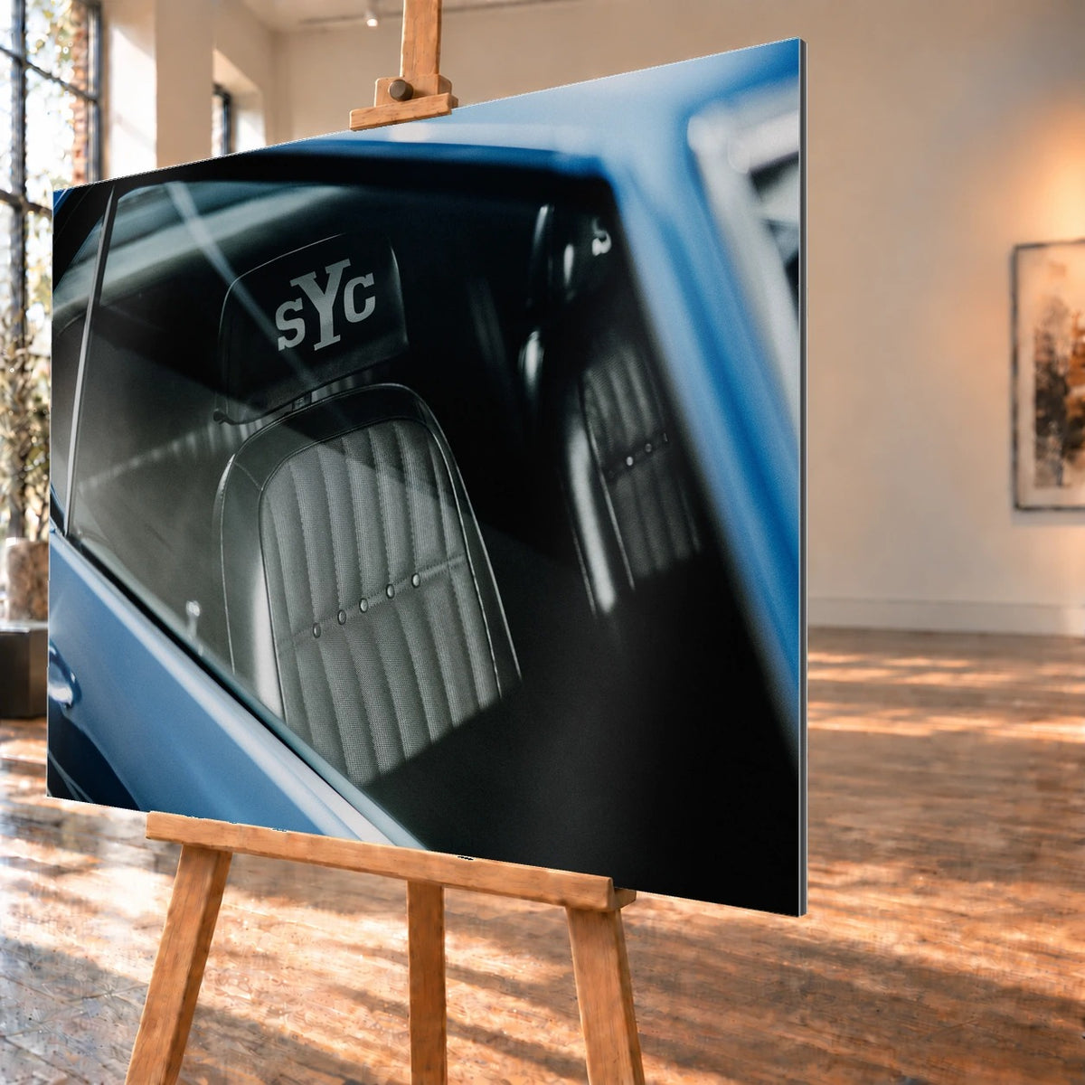 Die Innenraum-Nahaufnahme des blauen Camaro Yenko zeigt den hellen Sitz mit SYC-Schriftzug, eingefasst von Fenster und Karosserie. Motiv „Hero Moment 2“ als Alu gebürstet-Wandbild, schräg von der Seite auf einer Holzstaffelei.