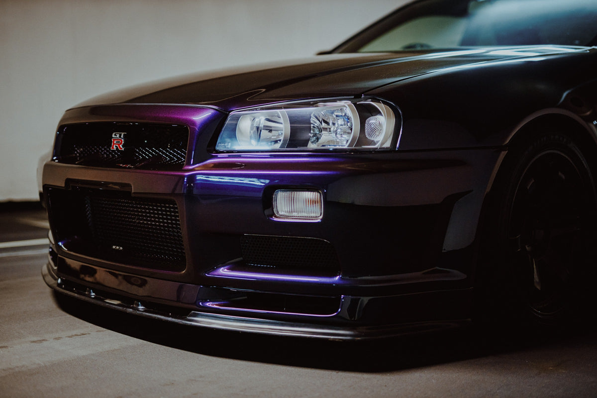 Die tiefe Nahaufnahme des Nissan Skyline R34 konzentriert sich auf Scheinwerfer, Grill und violette Frontschürze. Originalmotiv „Skyline Motion 2“ im Querformat.