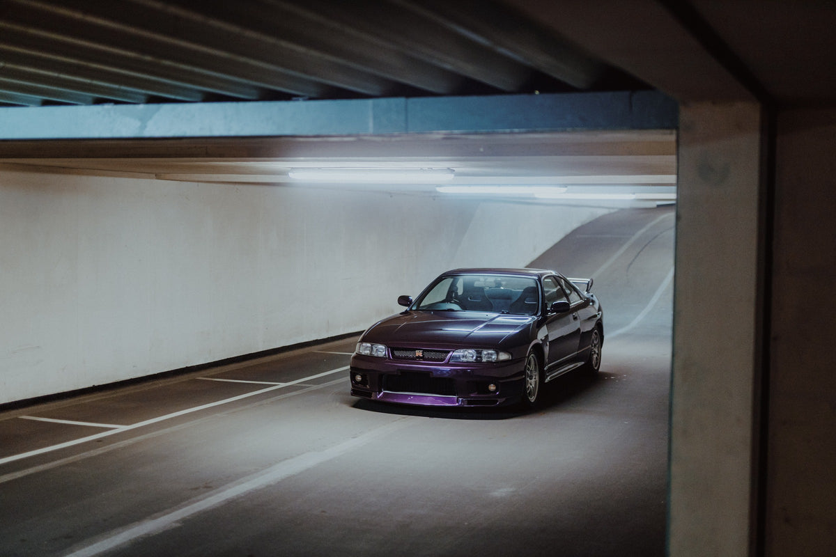 Der Nissan Skyline R33 ist schräg von vorn zwischen Betonstützen aufgenommen; violette Lackreflexe und kühles Deckenlicht bestimmen die Szene. Originalmotiv „Skyline Motion“ im Querformat.