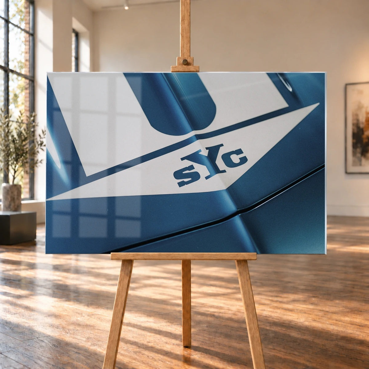 Die Nahaufnahme zeigt weiße Haubenstreifen und großen SYC-Schriftzug auf dem blauen Camaro Yenko. Motiv „Urban Light 2“ als Acrylglas-Wandbild, auf einer Holzstaffelei in einem hellen Galerieraum.