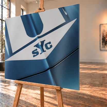 Die Nahaufnahme zeigt weiße Haubenstreifen und großen SYC-Schriftzug auf dem blauen Camaro Yenko. Motiv „Urban Light 2“ als Alu gebürstet-Wandbild, schräg von der Seite auf einer Holzstaffelei.