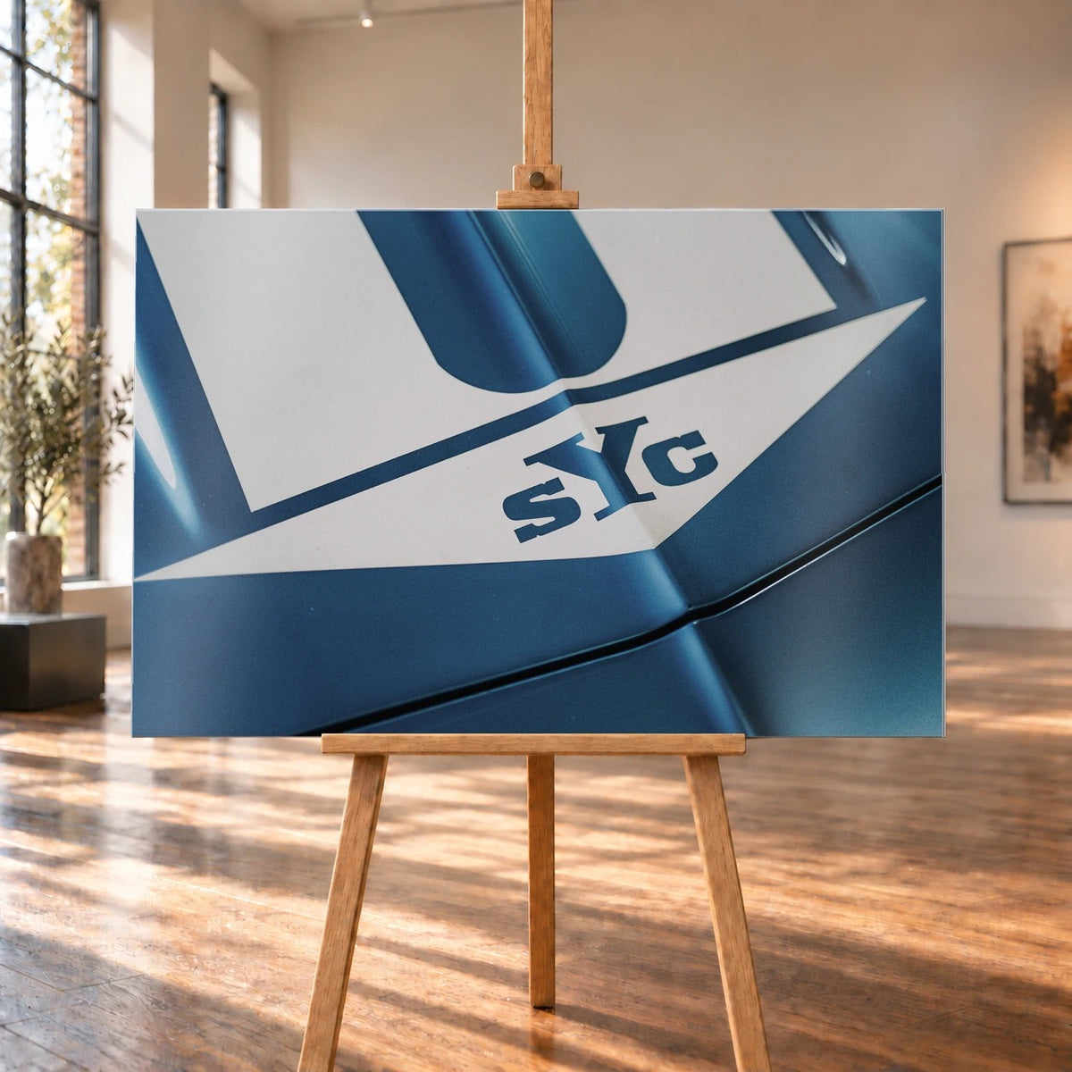 Die Nahaufnahme zeigt weiße Haubenstreifen und großen SYC-Schriftzug auf dem blauen Camaro Yenko. Motiv „Urban Light 2“ als Alu gebürstet-Wandbild, auf einer Holzstaffelei in einem hellen Galerieraum.