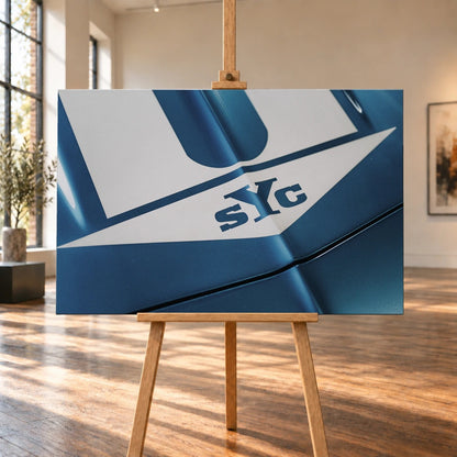 Die Nahaufnahme zeigt weiße Haubenstreifen und großen SYC-Schriftzug auf dem blauen Camaro Yenko. Motiv „Urban Light 2“ als Leinwand-Wandbild, auf einer Holzstaffelei in einem hellen Galerieraum.