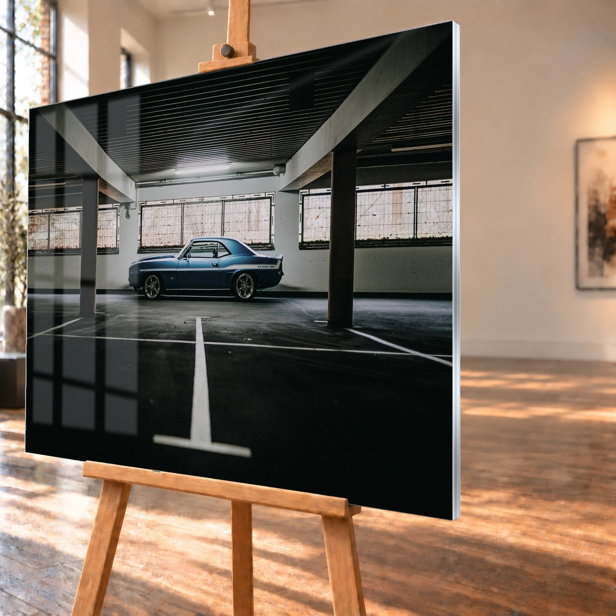 Der Camaro Yenko steht klein im Seitenprofil in einer großen, dunklen Industriehalle; helle Bodenmarkierung und Fensterlicht rahmen ihn ein. Motiv „Road Line“ als Acrylglas-Wandbild, schräg von der Seite auf einer Holzstaffelei.