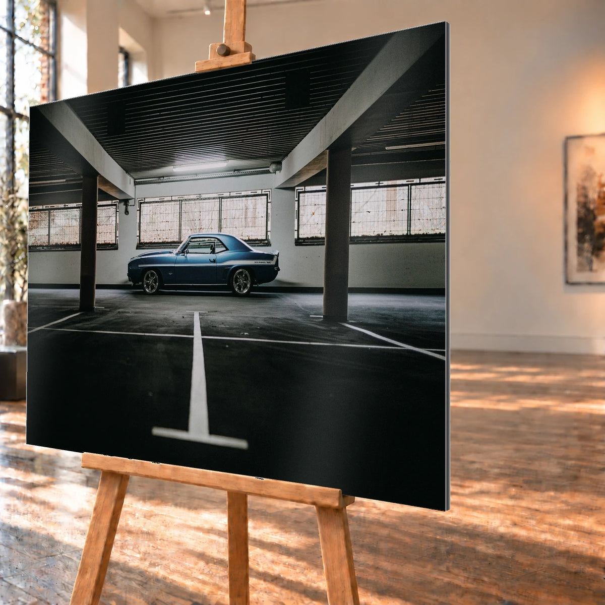 Der Camaro Yenko steht klein im Seitenprofil in einer großen, dunklen Industriehalle; helle Bodenmarkierung und Fensterlicht rahmen ihn ein. Motiv „Road Line“ als Alu gebürstet-Wandbild, schräg von der Seite auf einer Holzstaffelei.