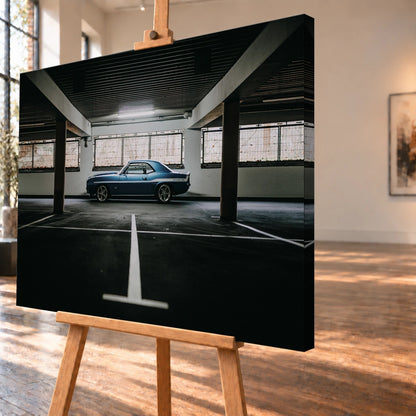 Der Camaro Yenko steht klein im Seitenprofil in einer großen, dunklen Industriehalle; helle Bodenmarkierung und Fensterlicht rahmen ihn ein. Motiv „Road Line“ als Leinwand-Wandbild, schräg von der Seite auf einer Holzstaffelei.