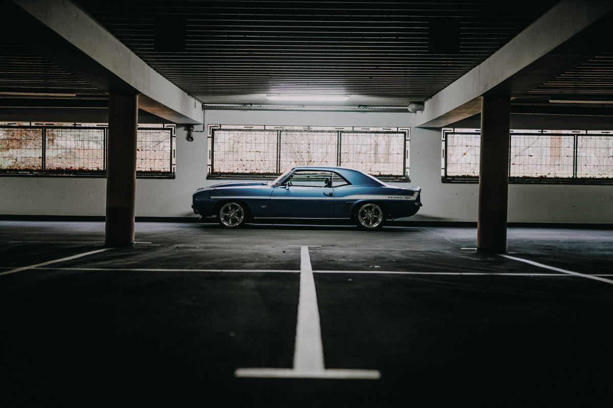 Der Camaro Yenko steht klein im Seitenprofil in einer großen, dunklen Industriehalle; helle Bodenmarkierung und Fensterlicht rahmen ihn ein. Originalmotiv „Road Line“ im Querformat.