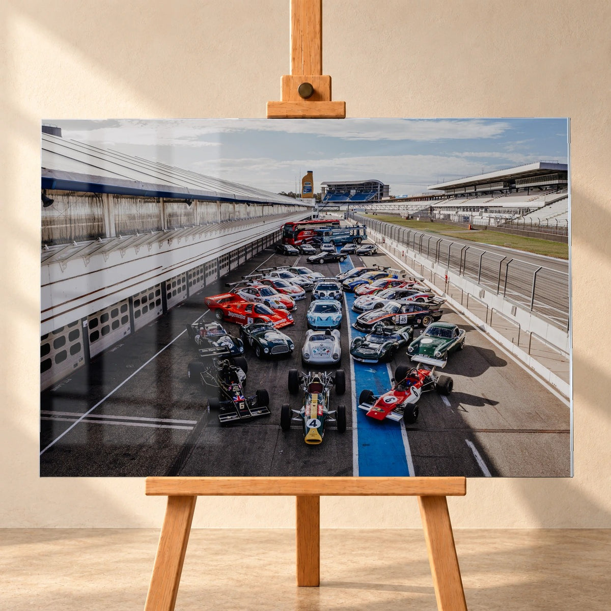 Eine große Gruppe unterschiedlicher Rennfahrzeuge steht im Fahrerlager zwischen Boxen und Rennstrecke; die erhöhte Perspektive zeigt die ganze Aufstellung. Motiv „Velocity Drive“ als Acrylglas-Wandbild, frontal auf einer Holzstaffelei.