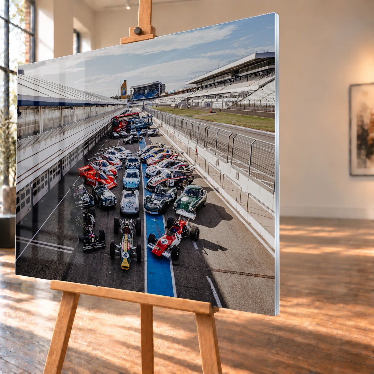 Eine große Gruppe unterschiedlicher Rennfahrzeuge steht im Fahrerlager zwischen Boxen und Rennstrecke; die erhöhte Perspektive zeigt die ganze Aufstellung. Motiv „Velocity Drive“ als Acrylglas-Wandbild, schräg von der Seite auf einer Holzstaffelei.