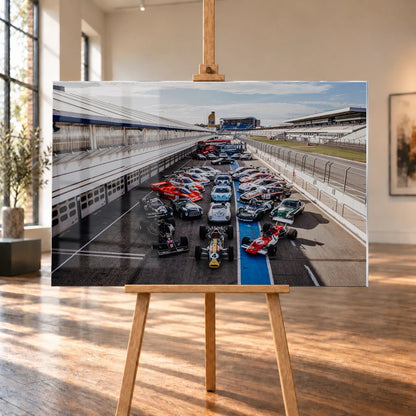 Eine große Gruppe unterschiedlicher Rennfahrzeuge steht im Fahrerlager zwischen Boxen und Rennstrecke; die erhöhte Perspektive zeigt die ganze Aufstellung. Motiv „Velocity Drive“ als Acrylglas-Wandbild, auf einer Holzstaffelei in einem hellen Galerieraum.