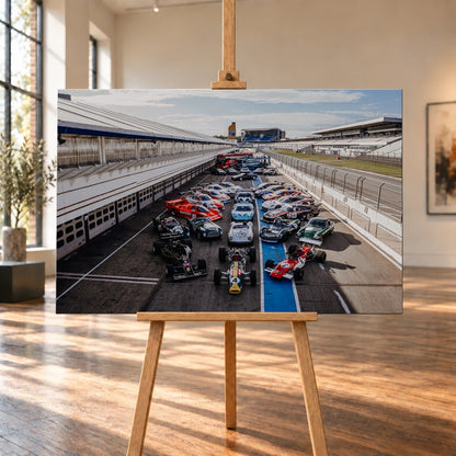 Eine große Gruppe unterschiedlicher Rennfahrzeuge steht im Fahrerlager zwischen Boxen und Rennstrecke; die erhöhte Perspektive zeigt die ganze Aufstellung. Motiv „Velocity Drive“ als Leinwand-Wandbild, auf einer Holzstaffelei in einem hellen Galerieraum.
