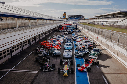 Eine große Gruppe unterschiedlicher Rennfahrzeuge steht im Fahrerlager zwischen Boxen und Rennstrecke; die erhöhte Perspektive zeigt die ganze Aufstellung. Originalmotiv „Velocity Drive“ im Querformat.