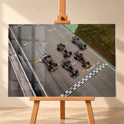 Fünf Lotus-Formelwagen stehen aus der Vogelperspektive nebeneinander auf der Startlinie; Markierungen und Asphalt erzeugen ein grafisches Raster. Motiv „Velocity Story“ als Acrylglas-Wandbild, frontal auf einer Holzstaffelei.