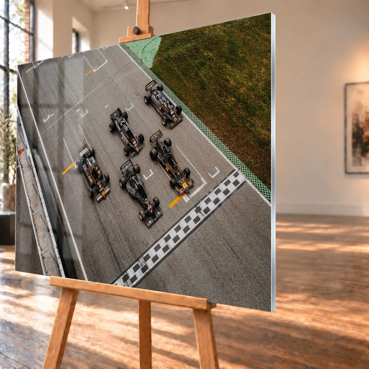 Fünf Lotus-Formelwagen stehen aus der Vogelperspektive nebeneinander auf der Startlinie; Markierungen und Asphalt erzeugen ein grafisches Raster. Motiv „Velocity Story“ als Acrylglas-Wandbild, schräg von der Seite auf einer Holzstaffelei.
