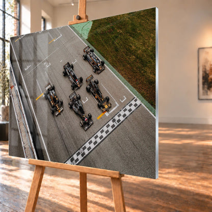 Fünf Lotus-Formelwagen stehen aus der Vogelperspektive nebeneinander auf der Startlinie; Markierungen und Asphalt erzeugen ein grafisches Raster. Motiv „Velocity Story“ als Acrylglas-Wandbild, schräg von der Seite auf einer Holzstaffelei.