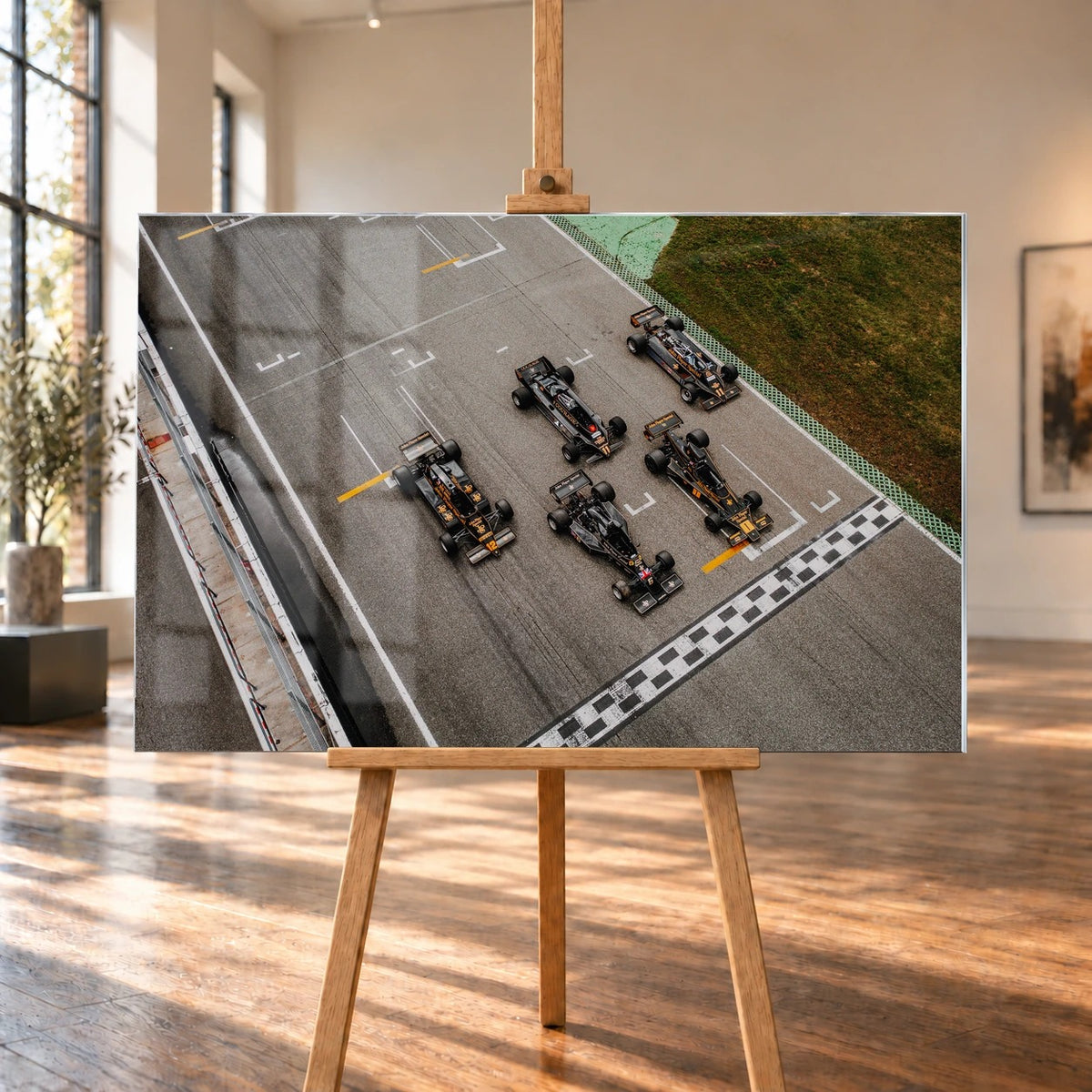 Fünf Lotus-Formelwagen stehen aus der Vogelperspektive nebeneinander auf der Startlinie; Markierungen und Asphalt erzeugen ein grafisches Raster. Motiv „Velocity Story“ als Acrylglas-Wandbild, auf einer Holzstaffelei in einem hellen Galerieraum.