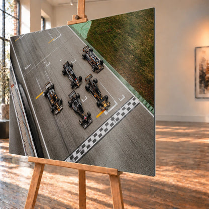 Fünf Lotus-Formelwagen stehen aus der Vogelperspektive nebeneinander auf der Startlinie; Markierungen und Asphalt erzeugen ein grafisches Raster. Motiv „Velocity Story“ als Alu gebürstet-Wandbild, schräg von der Seite auf einer Holzstaffelei.