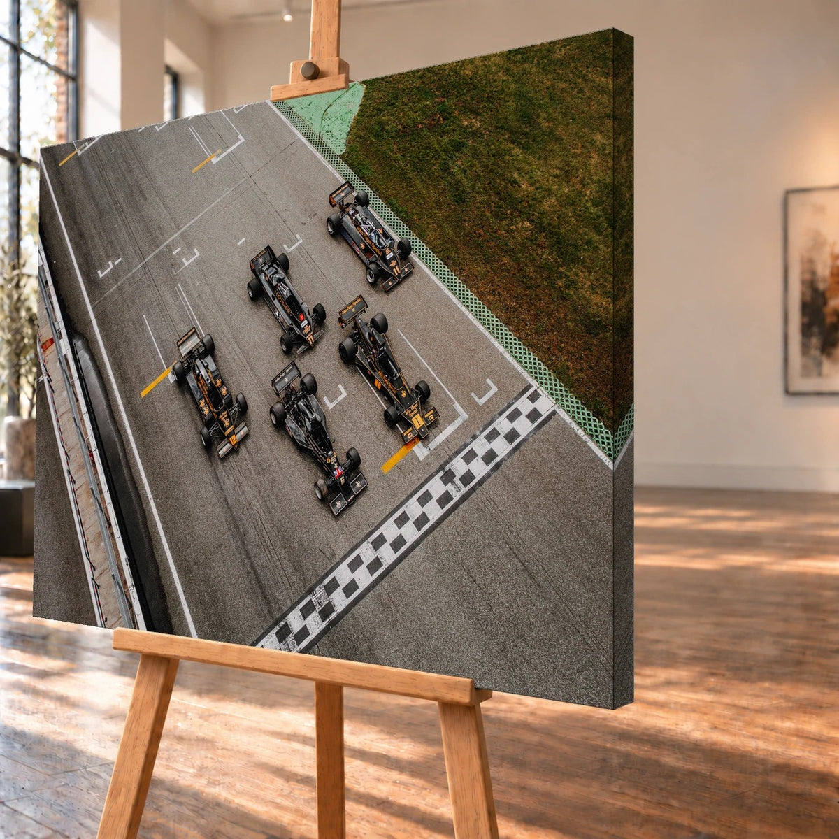 Fünf Lotus-Formelwagen stehen aus der Vogelperspektive nebeneinander auf der Startlinie; Markierungen und Asphalt erzeugen ein grafisches Raster. Motiv „Velocity Story“ als Leinwand-Wandbild, schräg von der Seite auf einer Holzstaffelei.