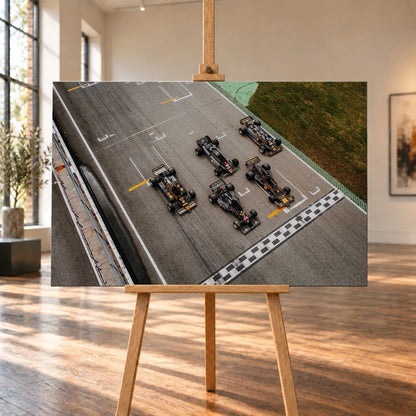 Fünf Lotus-Formelwagen stehen aus der Vogelperspektive nebeneinander auf der Startlinie; Markierungen und Asphalt erzeugen ein grafisches Raster. Motiv „Velocity Story“ als Leinwand-Wandbild, auf einer Holzstaffelei in einem hellen Galerieraum.