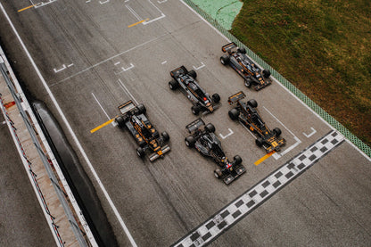 Fünf Lotus-Formelwagen stehen aus der Vogelperspektive nebeneinander auf der Startlinie; Markierungen und Asphalt erzeugen ein grafisches Raster. Originalmotiv „Velocity Story“ im Querformat.