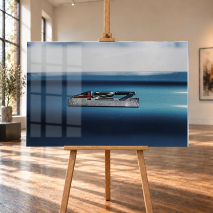 Das 427-Emblem ist als kleine Chrommarke auf der blauen Karosserie isoliert; die geringe Tiefenschärfe macht den Schriftzug zum Fokus. Motiv „Screen Pulse“ als Acrylglas-Wandbild, auf einer Holzstaffelei in einem hellen Galerieraum.