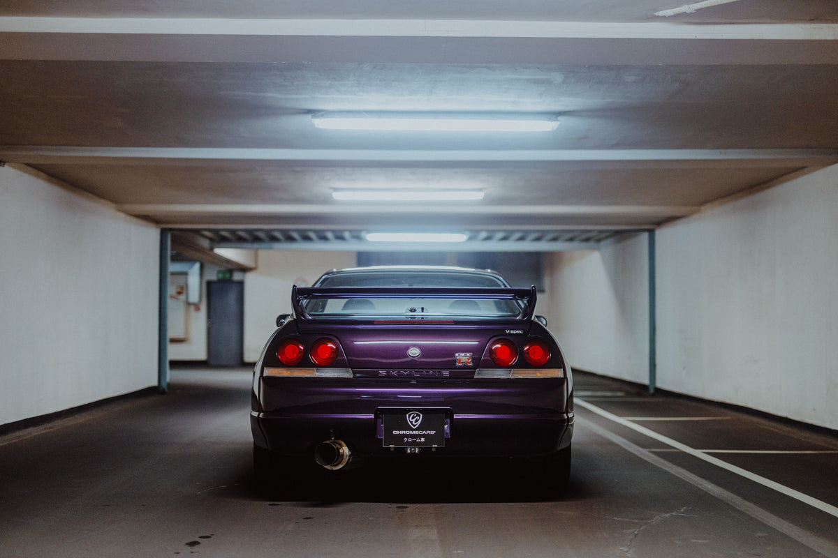 Der Nissan Skyline R33 steht mittig von hinten in einer leeren Parkhausachse; seine vier Rundlichter leuchten vor kühlem Beton. Originalmotiv „Midnight Depth 2“ im Querformat.