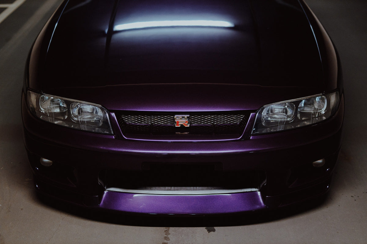 Eine enge Frontalansicht zeigt Grill, GT-R-Emblem und den violetten Frontsplitter des Nissan Skyline R33 vor dunklem Parkhausboden. Originalmotiv „Midnight Rhythm“ im Querformat.