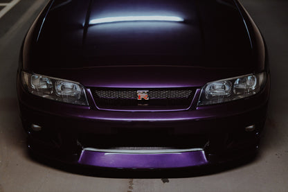 Eine enge Frontalansicht zeigt Grill, GT-R-Emblem und den violetten Frontsplitter des Nissan Skyline R33 vor dunklem Parkhausboden. Originalmotiv „Midnight Rhythm“ im Querformat.