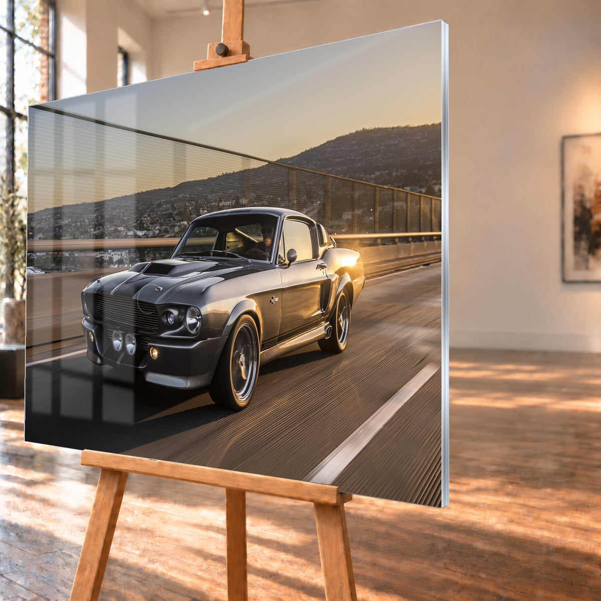 Grauer Eleanor-Sportwagen fährt im Abendlicht über eine Brücke, mit Stadt und Hügeln im Hintergrund; Visualisierung als Acrylglas-Wandbild, schräg von der Seite auf einer Holzstaffelei.