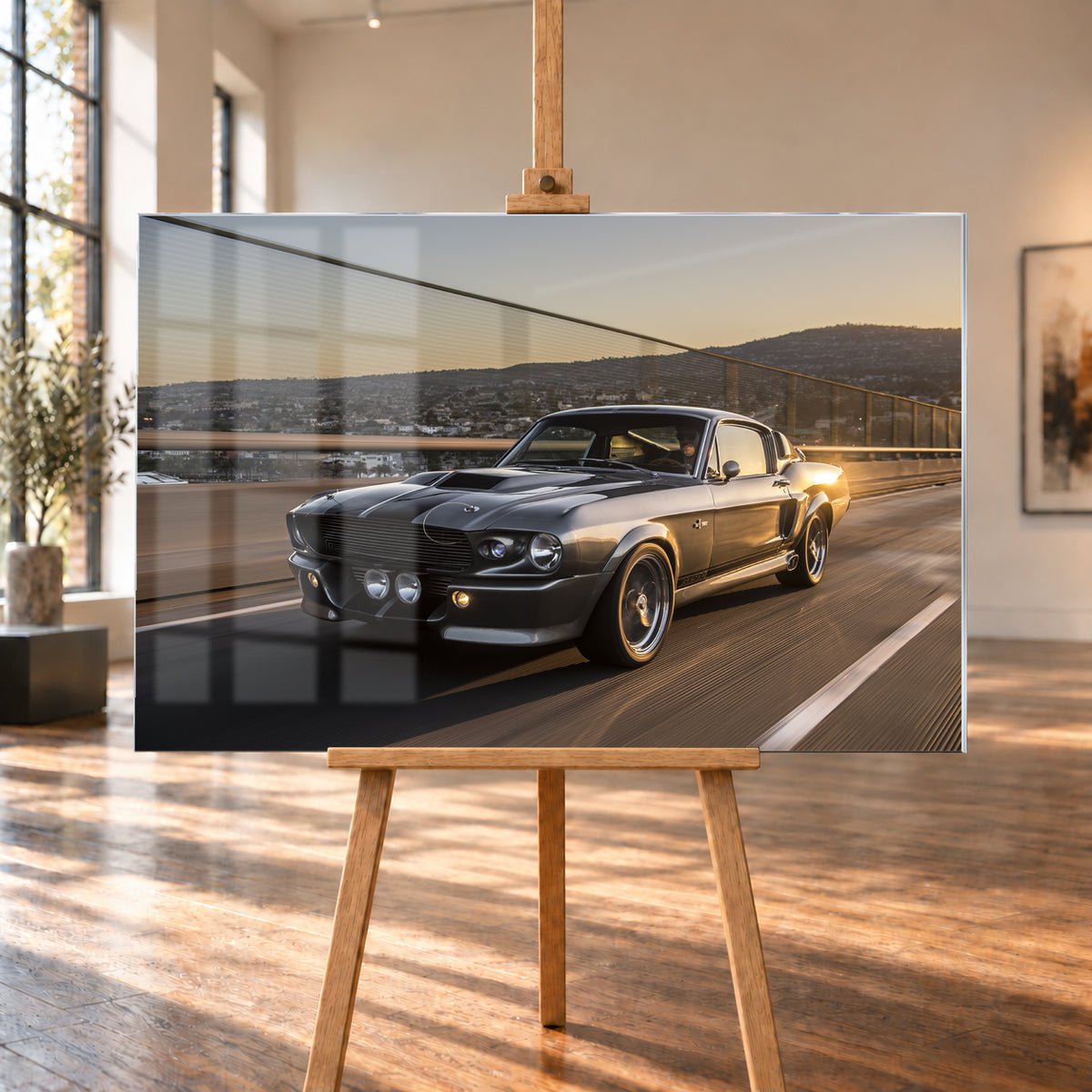 Grauer Eleanor-Sportwagen fährt im Abendlicht über eine Brücke, mit Stadt und Hügeln im Hintergrund; Visualisierung als Acrylglas-Wandbild, auf einer Holzstaffelei in einem hellen Galerieraum.