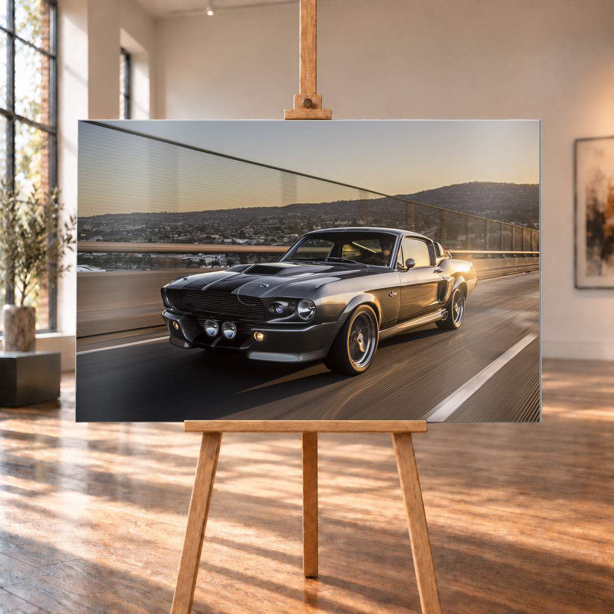 Grauer Eleanor-Sportwagen fährt im Abendlicht über eine Brücke, mit Stadt und Hügeln im Hintergrund; Visualisierung als Alu gebürstet-Wandbild, auf einer Holzstaffelei in einem hellen Galerieraum.