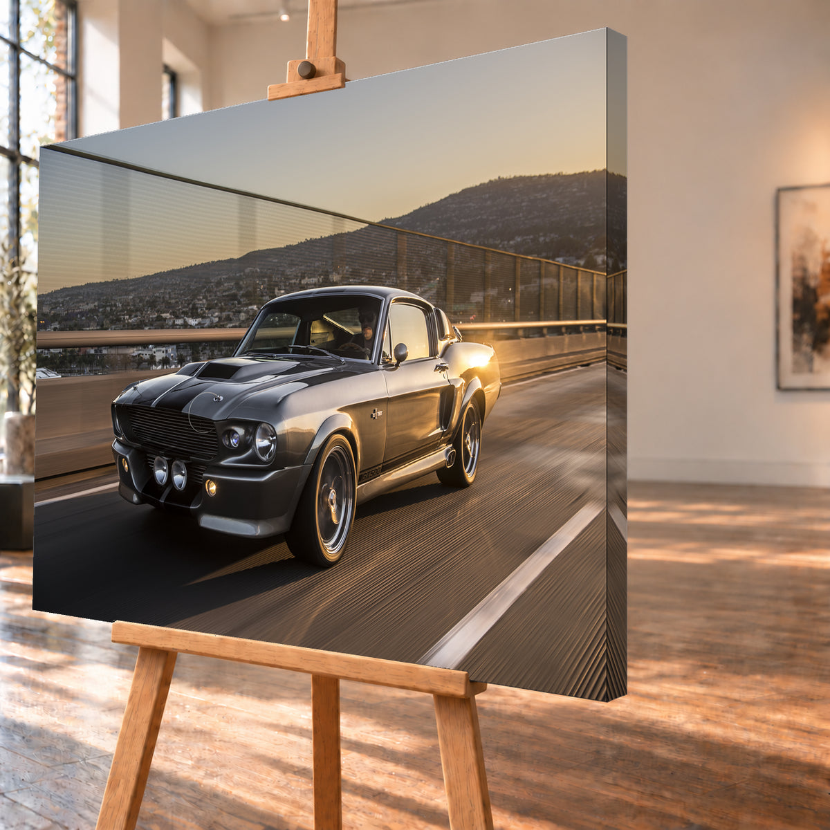 Grauer Eleanor-Sportwagen fährt im Abendlicht über eine Brücke, mit Stadt und Hügeln im Hintergrund; Visualisierung als Leinwand-Wandbild, schräg von der Seite auf einer Holzstaffelei.