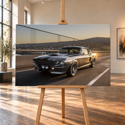 Grauer Eleanor-Sportwagen fährt im Abendlicht über eine Brücke, mit Stadt und Hügeln im Hintergrund; Visualisierung als Leinwand-Wandbild, auf einer Holzstaffelei in einem hellen Galerieraum.