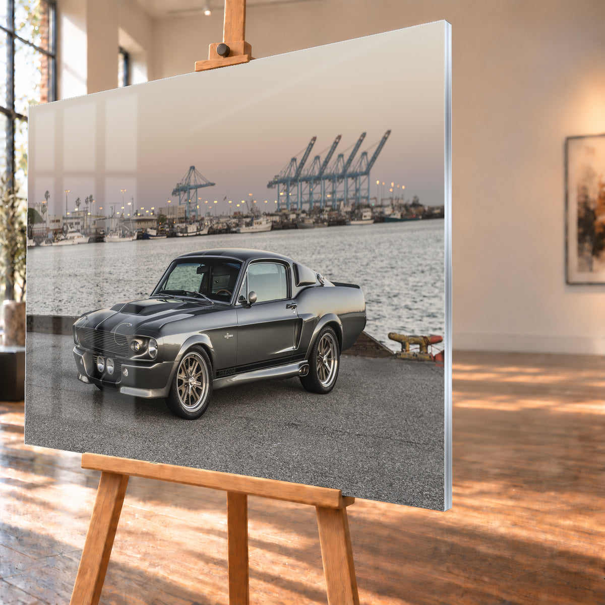 Dunkelgrauer Ford Mustang Shelby GT500 mit kontrastierenden Längsstreifen in Front-Dreiviertelansicht auf einer Hafenfläche; dahinter Wasser, Boote, Containerkräne, leuchtende Hafenlampen und ein pastellfarbener Abendhimmel; Visualisierung als Acrylglas-Wandbild, schräg von der Seite auf einer Holzstaffelei.