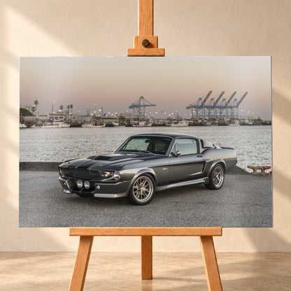 Dunkelgrauer Ford Mustang Shelby GT500 mit kontrastierenden Längsstreifen in Front-Dreiviertelansicht auf einer Hafenfläche; dahinter Wasser, Boote, Containerkräne, leuchtende Hafenlampen und ein pastellfarbener Abendhimmel; Visualisierung als Alu gebürstet-Wandbild, frontal auf einer Holzstaffelei.