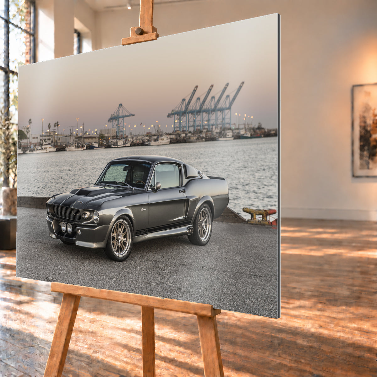 Dunkelgrauer Ford Mustang Shelby GT500 mit kontrastierenden Längsstreifen in Front-Dreiviertelansicht auf einer Hafenfläche; dahinter Wasser, Boote, Containerkräne, leuchtende Hafenlampen und ein pastellfarbener Abendhimmel; Visualisierung als Alu gebürstet-Wandbild, schräg von der Seite auf einer Holzstaffelei.