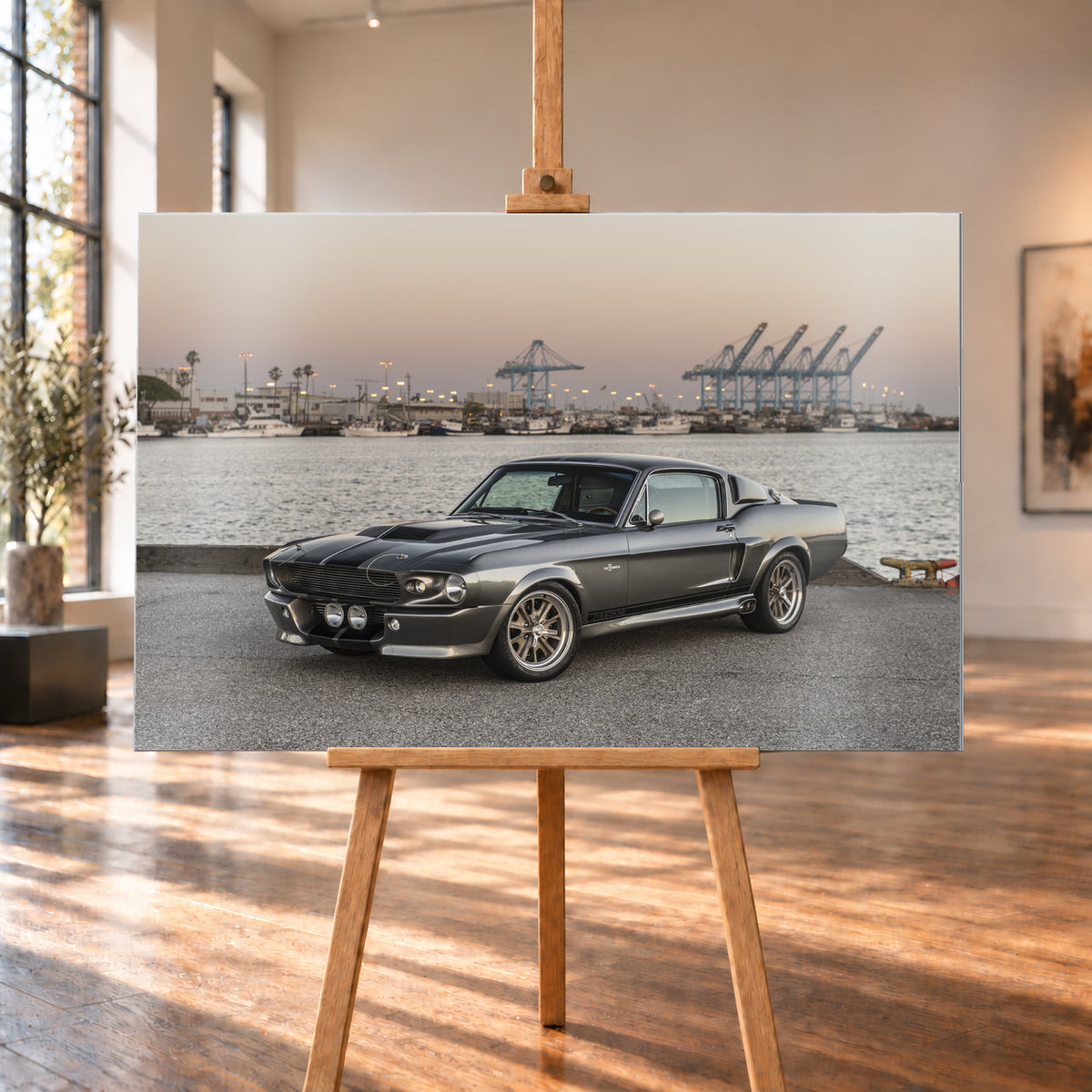 Dunkelgrauer Ford Mustang Shelby GT500 mit kontrastierenden Längsstreifen in Front-Dreiviertelansicht auf einer Hafenfläche; dahinter Wasser, Boote, Containerkräne, leuchtende Hafenlampen und ein pastellfarbener Abendhimmel; Visualisierung als Alu gebürstet-Wandbild, auf einer Holzstaffelei in einem hellen Galerieraum.