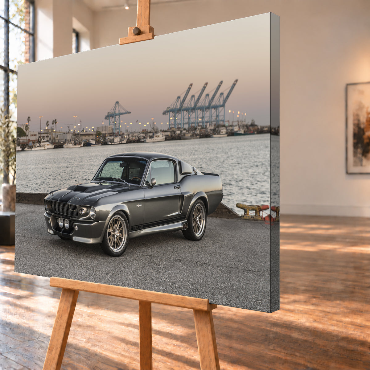 Dunkelgrauer Ford Mustang Shelby GT500 mit kontrastierenden Längsstreifen in Front-Dreiviertelansicht auf einer Hafenfläche; dahinter Wasser, Boote, Containerkräne, leuchtende Hafenlampen und ein pastellfarbener Abendhimmel; Visualisierung als Leinwand-Wandbild, schräg von der Seite auf einer Holzstaffelei.