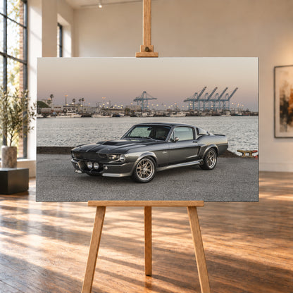 Dunkelgrauer Ford Mustang Shelby GT500 mit kontrastierenden Längsstreifen in Front-Dreiviertelansicht auf einer Hafenfläche; dahinter Wasser, Boote, Containerkräne, leuchtende Hafenlampen und ein pastellfarbener Abendhimmel; Visualisierung als Leinwand-Wandbild, auf einer Holzstaffelei in einem hellen Galerieraum.
