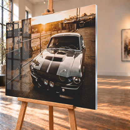 Silbergraues klassisches Fastback mit schwarzen Längsstreifen, schräg von vorn fotografiert, vor Maschendrahtzaun und Tor auf einem Industriegelände bei tief stehender, warm leuchtender Sonne; Visualisierung als Acrylglas-Wandbild, schräg von der Seite auf einer Holzstaffelei.