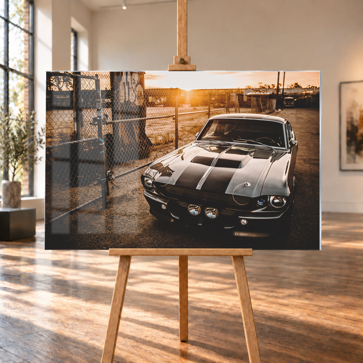 Silbergraues klassisches Fastback mit schwarzen Längsstreifen, schräg von vorn fotografiert, vor Maschendrahtzaun und Tor auf einem Industriegelände bei tief stehender, warm leuchtender Sonne; Visualisierung als Acrylglas-Wandbild, auf einer Holzstaffelei in einem hellen Galerieraum.