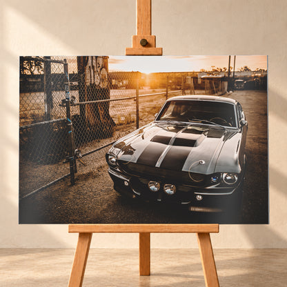 Silbergraues klassisches Fastback mit schwarzen Längsstreifen, schräg von vorn fotografiert, vor Maschendrahtzaun und Tor auf einem Industriegelände bei tief stehender, warm leuchtender Sonne; Visualisierung als Alu gebürstet-Wandbild, frontal auf einer Holzstaffelei.
