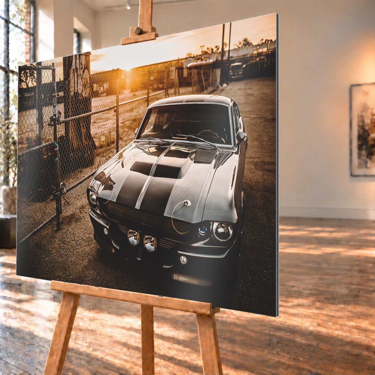 Silbergraues klassisches Fastback mit schwarzen Längsstreifen, schräg von vorn fotografiert, vor Maschendrahtzaun und Tor auf einem Industriegelände bei tief stehender, warm leuchtender Sonne; Visualisierung als Alu gebürstet-Wandbild, schräg von der Seite auf einer Holzstaffelei.