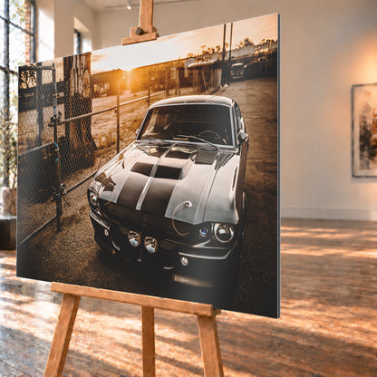 Silbergraues klassisches Fastback mit schwarzen Längsstreifen, schräg von vorn fotografiert, vor Maschendrahtzaun und Tor auf einem Industriegelände bei tief stehender, warm leuchtender Sonne; Visualisierung als Alu gebürstet-Wandbild, schräg von der Seite auf einer Holzstaffelei.
